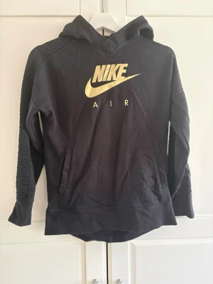 Svart Nike Air hoodie med gul logga - Snygg svart hoodie från Nike med stor guldfärgad Nike Air-logga på bröstet. Hoodien har huva, magficka och coola detaljer med upphöjt mönster längs ärmarna. Perfekt för en sportig och avslappnad stil.