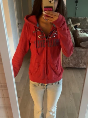 Röd zip hoodie från Hollister - Säljer en röd hoodie från Hollister med dragkedja, huva och fickor framtill. Hoodien har blå textdetaljer på bröstet och snören i huvan. Perfekt för en avslappnad och cool stil. Materialet är mjukt och skönt.