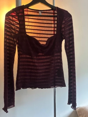 Mörklila mesh topp med lång ärm - Säljer en mörklila mesh topp med långa ärmar och transparent randigt mönster. Toppen har en markerad byst med twistad detalj framtill och är figurnära i modellen. Perfekt för dig som vill ha en edgy och trendig look. Passar även S