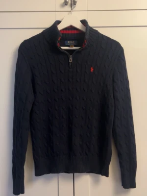 Mörkblå kabelstickad tröja Polo Ralph Lauren - Mörkblå kabelstickad tröja från Polo Ralph Lauren med halv dragkedja och hög krage. Tröjan har ett rött broderat logomärke på bröstet och ribbade muddar vid ärmslut och nederkant. Perfekt för dig som gillar klassisk och stilren look.