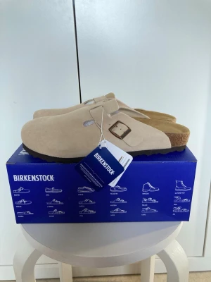 Beige Birkenstock tofflor mocka - Säljer ett par snygga beige Birkenstock tofflor i mocka med klassisk korksula och justerbart spänne i metall. Perfekta för dig som vill ha både stil och komfort. Levereras i originalkartong med taggar kvar.