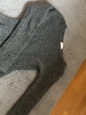 H&M tröja -  mörkgrå stickad tröja med peplumskärning och långa ärmar. Tröjan har en rund halsringning och är tillverkad i ett mjukt, stretchigt material som ger en skön passform. Perfekt för dig som gillar en feminin siluett och vill ha något extra till din outfit.