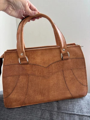 Retro Brun handväska i läder från Pragt Design - Snygg retro handväska i brunt läder från Pragt Design från 90-talet . Väskan har två stadiga handtag, dekorativa sömmar och en dragkedja upptill. Klassisk rektangulär form och stilren design som passar till många outfits. Mått 34x22x10 cm 