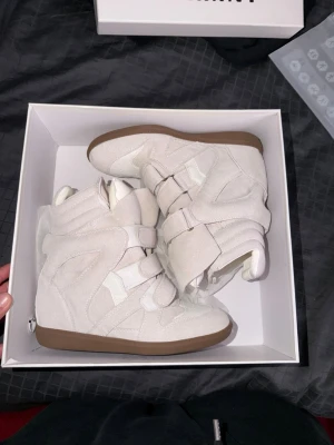 Isabel Marant  - Säljer ett par vita höga sneakers med chunky design och flera breda kardborreband. Skorna har en cool siluett med vadderad häl och ovandel i mockaliknande material. Yttersulan är i gummi och kontrasterar snyggt mot den ljusa ovandelen. köpta på plick av någon annan!