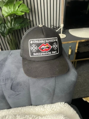 Svart truckerkeps med brodyr - Svart truckerkeps med nät på sidorna och broderad patch framtill med texten 'Chrome Hearts Hollywood USA', blommor och röda läppar. Justerbar baktill för perfekt passform. Perfekt accessoar för en streetwear-look.