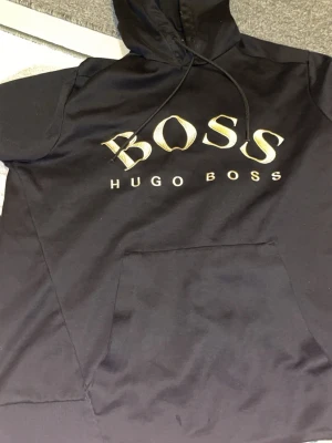 Svart hoodie från Hugo Boss - Svart hoodie med stor BOSS-logga i guld på bröstet och texten HUGO BOSS under. Klassisk huva med snören och en stor magficka framtill. Perfekt för dig som gillar stilrena och exklusiva plagg.