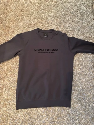 Mörkgrå sweatshirt från Armani Exchange - Snygg mörkgrå sweatshirt från Armani Exchange med tryckt logga och texten 'Milano/New York' på bröstet. Klassisk rund halsringning och långa ärmar. Perfekt för en avslappnad och trendig look.