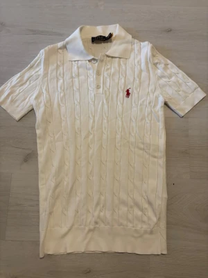 Vit kabelstickad pikétröja Polo Ralph Lauren - Vit kabelstickad pikétröja från Polo Ralph Lauren med korta ärmar och klassisk krage. Tröjan har knappar framtill och den ikoniska röda logotypen broderad på bröstet. Perfekt för en clean och stilren look.