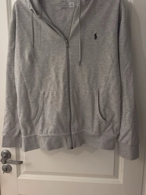 Grå zip-hoodie från Polo Ralph Lauren - Snygg grå hoodie med dragkedja från Polo Ralph Lauren. Klassisk design med huva, två fickor framtill och broderad logga på bröstet. Perfekt för en avslappnad och stilren look. Mjuk bomullsblandning och ribbade muddar.