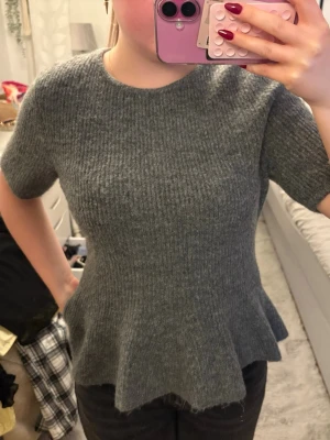 Grå stickad topp med volang - Grå stickad topp med korta ärmar och rund halsringning. Modellen har en figurnära passform upptill och en volangkant nedtill som ger en snygg siluett. Perfekt att styla med jeans eller kjol för en trendig look.