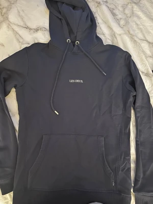 Mörkblå hoodie från Les Deux - Snygg mörkblå hoodie från Les Deux med broderad logga på bröstet. Klassisk känguruficka framtill, justerbar huva med dragsko och ribbade muddar. Tillverkad i mjukt bomullsmaterial som är skönt mot huden.