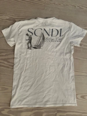 Vit SCNDI Sailing Club t-shirt - Vit t-shirt med tryck från SCNDI Sailing Club Stockholm. Framsidan har en liten logga på bröstet och baksidan har ett stort motiv med segelbåt och person. Klassisk rund hals och korta ärmar. Perfekt för en avslappnad och stilren look.