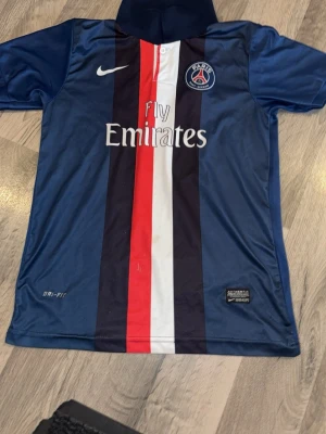 PSG matchtröja blå Nike - Snygg blå PSG matchtröja från Nike med vertikala ränder i mörkblått, vitt och rött. Tröjan har Paris Saint-Germain klubbmärke på bröstet och Fly Emirates-tryck framtill. Tillverkad i lätt och snabbtorkande Dri-Fit material, perfekt för fotbollsträning.