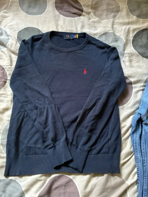 Mörkblå stickad tröja Polo Ralph Lauren - Mörkblå stickad tröja från Polo Ralph Lauren med rund halsringning och röd broderad logga på bröstet. Tröjan har långa ärmar och ribbade muddar vid ärmslut och nederkant. Perfekt för lager på lager och stilren look.