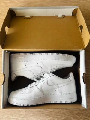 Nike Air Force 1 - vita sneakers - Original, köpta på zalando.se, men aldrig använda. Helt nya oanvända helvita Nike Air Force 1 sneakers i lågt utförande. Skorna har en stilren design med perforerade detaljer på tån, kraftig sula och ikoniska swoosh-loggan på sidan. Tillverkade i slätt läder med vita skosnören och diskret AIR-text på sulan.