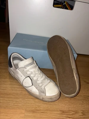 Vita Philippe Model sneakers - Snygga vita sneakers från Philippe Model med grå detaljer och svart häl. Skorna har en klassisk låg siluett, vita snören och en robust gummisula. Materialet är en mix av läder och mocka för en cool och avslappnad look.
