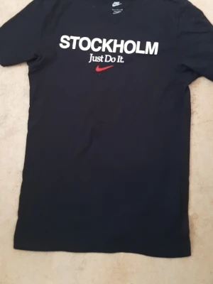 Svart Nike t-shirt Stockholm S - Säljer en svart t-shirt från Nike med texten 'STOCKHOLM Just Do It.' och en röd swoosh på bröstet. Klassisk passform och rund halsringning. Perfekt för dig som gillar streetwear och vill ha något stilrent med en touch av sport.