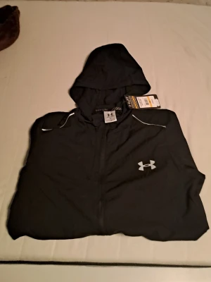 Svart vindjacka från Under Armour - Svart vindjacka från Under Armour med huva och hel dragkedja. Jackan har vita detaljer vid axlarna, logga på bröstet och texten 'STORM' på ärmen. Lätt material som passar perfekt för blåsiga dagar.