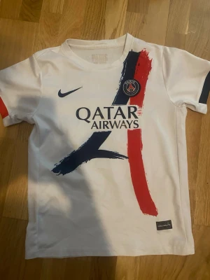 PSG matchtröja Vitinha 17 Nike - Säljer en vit PSG matchtröja från Nike med tryck Vitinha 17 på ryggen. Tröjan har blå och röda penseldrag på framsidan, PSG-logga på bröstet och Qatar Airways som sponsor. Korta ärmar med detaljer i rött och blått. Perfekt för fotbollsfans!