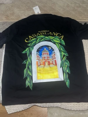 Svart Avenida Casablanca t-shirt - Svart t-shirt med färgglad tryckt grafik på ryggen och mindre tryck på bröstet. Motivet föreställer en fönsterbåge med gröna blad och en byggnad i bakgrunden, samt texten 'Avenida Casablanca'. Rund hals och korta ärmar.