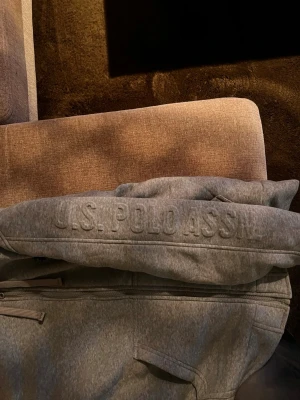 Grå hoodie från U.S. Polo Assn. - Grå hoodie med dragkedja från U.S. Polo Assn. Broderad logga på bröstet och text på ärmen. Utrustad med huva och dragsko samt två fickor framtill. Perfekt för en kall kväll eftersom det är lurv innuti 