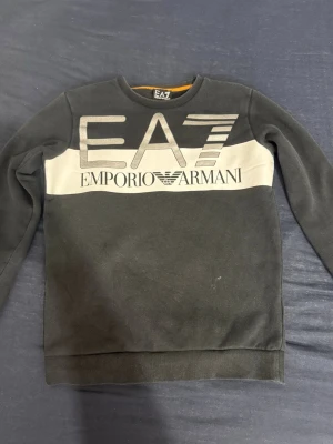 Svart tröja från Emporio Armani - Snygg svart tröja från Emporio Armani med stor EA7-logga och vit panel över bröstet. Tröjan har rund halsringning och långa ärmar. Perfekt för dig som gillar stilrena och exklusiva plagg.