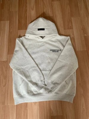Essentials Fear of God hoodie - Säljer en ljusgrå hoodie från Essentials Fear of God med svart logga tryckt på bröstet. Hoodien har en klassisk huva och ribbade muddar vid ärmar och nederkant. Perfekt för dig som gillar streetwear och enkel stil. Oanvänd men kommer utan prislapp.