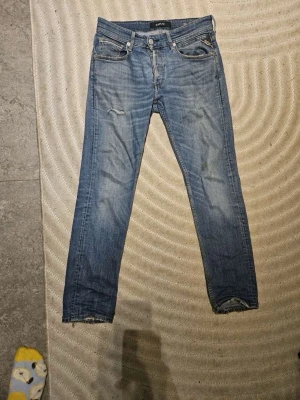 Ljusblå Replay jeans med slitningar - Säljer ett par ljusblå replay jeans med raka ben och slitningar på låret och vid benslut. Jeansen har klassisk femficksmodell och gylf med dragkedja. Perfekta för en avslappnad och trendig look.