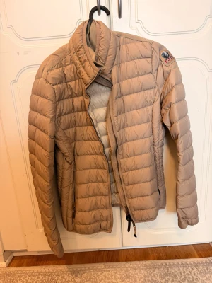 Beige dunjacka från Parajumpers - Säljer en beige dunjacka från Parajumpers med klassisk quiltad design och hög krage. Jackan har dragkedja framtill, två sidofickor och en patch med PJS-logga på ärmen. Perfekt för kalla vinterdagar och riktigt snygg till alla outfits.