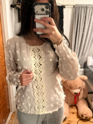 Beige blus med spetsdetaljer - Söt beige blus med långärmad design och transparent tyg dekorerat med små bollar. Framsidan har en bred spetsdetalj i vitt och ärmarna har smockade muddar. Perfekt för dig som gillar romantisk och bohemisk stil.