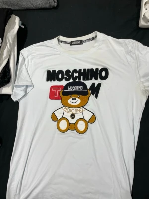 Vit Moschino T-shirt med björn - Säljer en vit T-shirt från Moschino med ett stort broderat motiv av en björn på framsidan. T-shirten har tryckt text och detaljer i svart, rött och gult. Perfekt för dig som gillar statement-plagg och lekfull design.