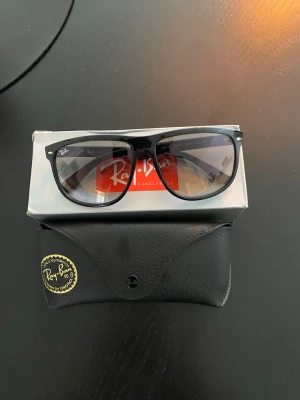 Svarta Ray-Ban Wayfarer solglasögon - Klassiska svarta Ray-Ban Wayfarer solglasögon med tidlös design och mörka glas. Bågarna är i plast och modellen har den ikoniska Ray-Ban-loggan på linsen. Perfekta för att höja din stil under soliga dagar. Fodral medföljer.