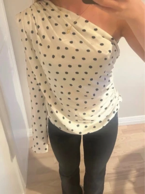Prickig oneshoulder topp - Säljer en vit oneshoulder topp med svarta prickar. Toppen har en lång ärm på ena sidan och är axelbandslös på andra sidan. Materialet känns mjukt och följsamt, perfekt för dig som vill sticka ut lite extra.