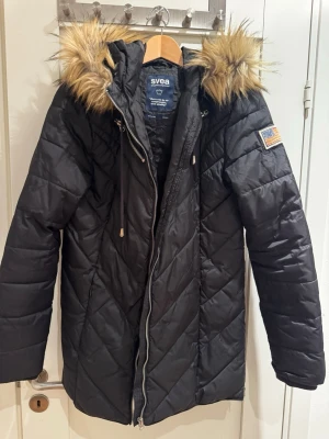 Svart dunjacka med fuskpäls från Svea - Snygg svart dunjacka från Svea med lång passform och quiltat mönster. Jackan har huva med beige fuskpäls, dragkedja framtill och en amerikansk flaggpatch på ärmen. Perfekt för kalla vinterdagar och riktigt varm.