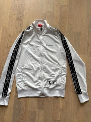 Vit zip hoodie från Champion - Snygg vit zip hoodie från Champion med svarta ränder och logga längs ärmarna. Använd men finns inga tecken på användning.