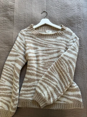 Beige zebramönstrad stickad tröja - Stickad tröja från Object i beige och vitt zebramönster. Tröjan har rund halsringning, långa ärmar och ribbade muddar vid ärmslut och nederkant. Perfekt för dig som gillar djurmönster och vill ha en mysig, trendig look. 