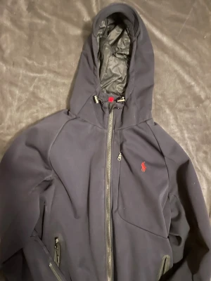 Ralph lauren jacka  - Snygg mörkblå hoodiejacka med hel dragkedja framtill och två sidofickor med dragkedja. Jackan har en broderad röd logga på bröstet och justerbar huva. Perfekt för kyliga dagar och enkel att matcha med olika outfits.
