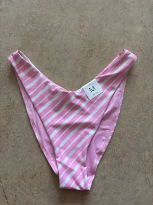 Rosa och vit randig bikiniunderdel - Säljer en bikiniunderdel i klassisk modell med diagonala rosa och vita ränder. Materialet är stretchigt och mjukt, perfekt för stranden eller poolen. Snyggt V-format snitt framtill för en trendig look.