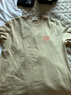 Beige Gallery Dept t-shirt - Snygg beige t-shirt från Gallery Dept. med tryck i rött fram och stort tryck på ryggen. Klassisk passform och rund halsringning. Perfekt för dig som gillar streetwear och enkel stil.