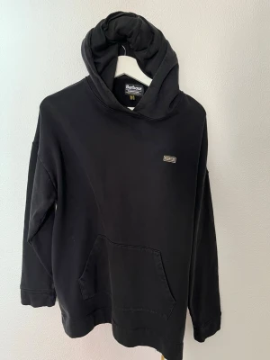 Svart hoodie från Barbour med dragkedja - Snygg svart hoodie från Barbour med huva och magficka. Har en guldfärgad dragkedja på sidan som cool detalj. Liten Barbour-logga på bröstet. Tillverkad i mjukt bomullsmaterial och har en avslappnad passform. Perfekt för en chill och stilren look.