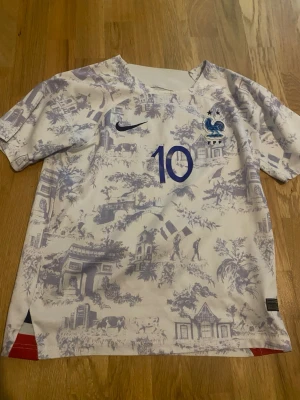 Frankrike fotbollströja Mbappé #10 - Snygg vit fotbollströja från Nike med blått tryck, nummer 10 och Mbappé på ryggen. Tröjan har franska landslagets emblem på bröstet och ett unikt blått mönster med franska landmärken. Perfekt för dig som älskar fotboll och Frankrike.