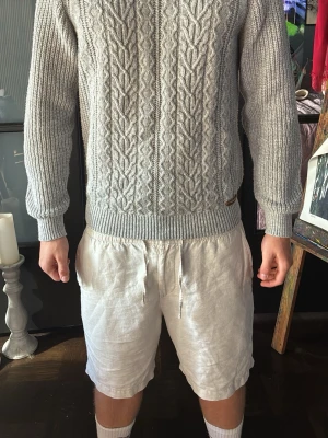 Ljusgrå kabelstickad tröja - Snygg ljusgrå kabelstickad tröja med klassiskt flätmönster framtill. Tröjan har rund halsringning, ribbade muddar och nederkant. Perfekt för lager på lager och passar till det mesta. Materialet ser ut att vara mjukt och bekvämt, troligen bomull eller ullblandning.