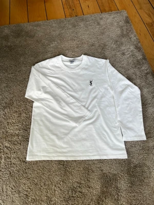 Vit långärmad topp med YSL-brodyr - Vit långärmad topp med rund hals och svart YSL-brodyr på bröstet. Tillverkad i mjuk bomull som känns skön mot huden. Enkel och clean design som passar till det mesta. Är storlek M men passar som S
