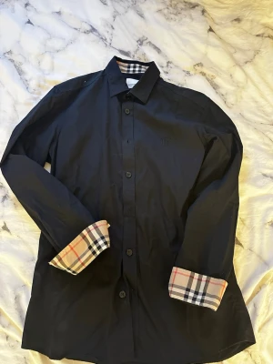 Svart skjorta från Burberry - Stilren svart skjorta från Burberry med klassiskt rutigt mönster på insidan av kragen och ärmsluten. Skjortan har knäppning framtill, lång ärm och diskret broderad logga på bröstet. Perfekt för dig som gillar exklusiva detaljer och tidlös design. Kvittot finns även att få. 