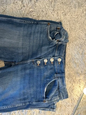 Blå raka jeans med knappar - Säljer ett par klassiska blå jeans med raka ben och synliga silverfärgade knappar i gylfen. Jeansen har fem fickor och en ljus tvättad look. Materialet är denim i bomull och passformen är avslappnad. Perfekta till vardagsstilen!