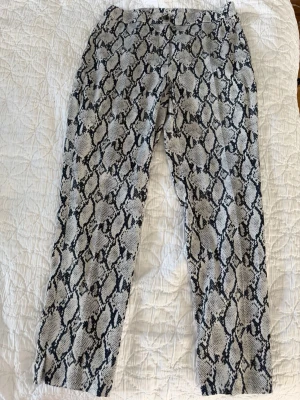 Chinos med ormmönster i grått och blått - Snygga chinos med trendigt ormmönster i grått och blått. Byxorna har rak passform och normal midja. Materialet känns mjukt och följsamt, perfekt för dig som vill sticka ut med din stil. Stängs med knapp och dragkedja framtill.