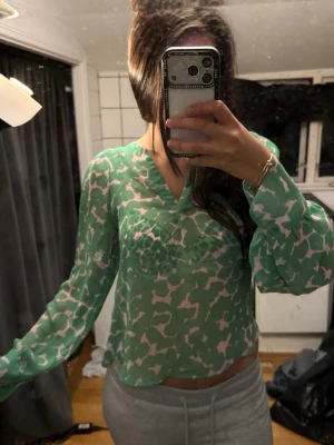 Grön transparent blus med mönster - Snygg grön blus i transparent material med ljusrosa abstrakt mönster. Blusen har v-ringning och långa, luftiga ärmar. Perfekt för dig som gillar en trendig och luftig stil.