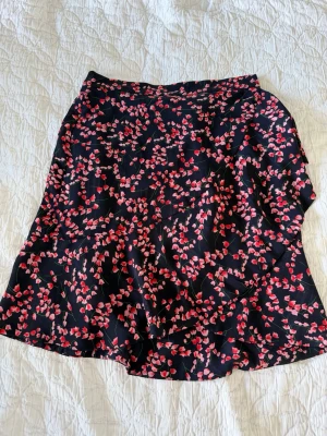 Mörkblå blommig omlottkjol - Snygg mörkblå omlottkjol  med små rosa och röda blommor. Kjolen har en lätt utställd form och är tillverkad i ett mjukt, tunt material som känns skönt mot huden. Perfekt för dig som gillar feminina och lekfulla plagg med fina detaljer.