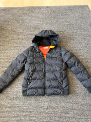 Parajumpers vinterjacka - Snygg svart dunjacka från Parajumpers med huva och dragkedja framtill. Jackan har två sidofickor med dragkedja och en patch på ärmen med Parajumpers-logga. Insidan är orange och jackan har ribbade muddar vid ärmslut och nederkant.