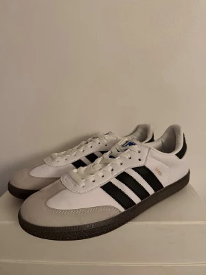 Adidas Samba vita och svarta sneakers - Klassiska Adidas Samba sneakers i vitt och svart med de ikoniska tre ränderna på sidan. Ovandelen är i läder och mocka, med brun gummisula och snörning. Perfekta för en sportig och stilren look. 44/45 helt nya inpincip 